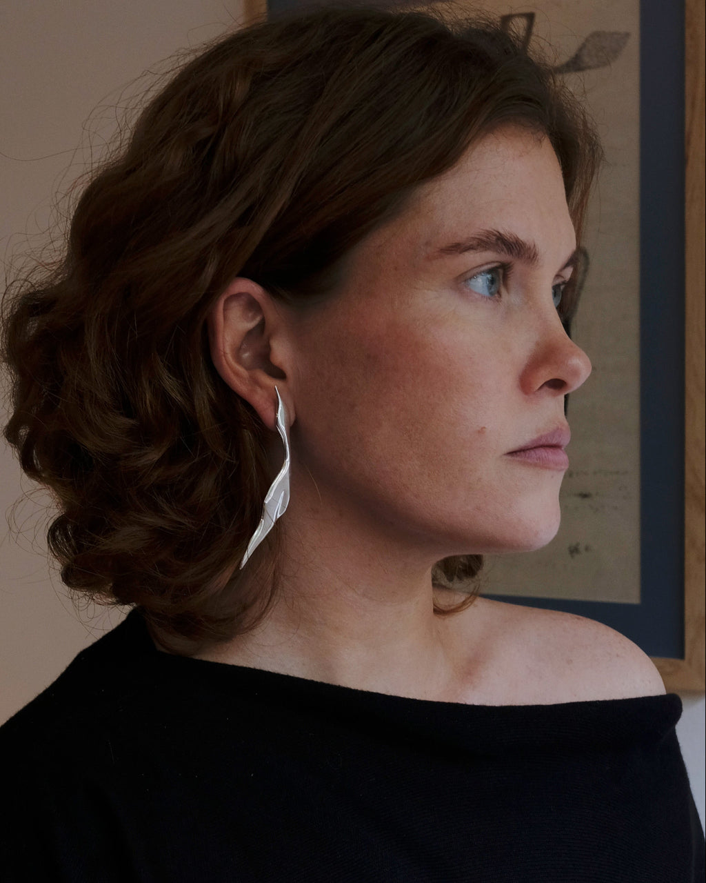 Woman in a black off-shoulder top standing in a room with a framed picture on the wall. wearing sterling silver Virginia Jirash earrings