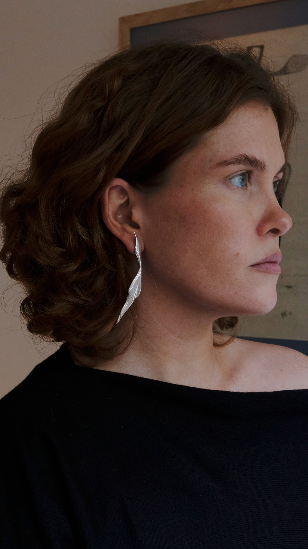 Woman in a black off-shoulder top standing in a room with a framed picture on the wall . Wearing Virginia Jirash Garzas silver earrings